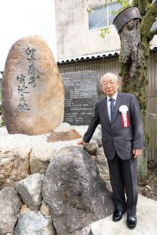 Hiroshi Doi Sensei in a suit with a ribbon stands beside large engraved memorial stones and rocks outdoors.