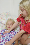 A young girl in a red shirt gently places her hands on another girl’s chest for a Reiki session.