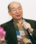 Hiroshi Doi smiling and holding a microphone while speaking at an event, wearing a patterned shirt and blazer.