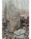 A large, upright natural stone monument placed in a wooded area covered with fallen autumn leaves.