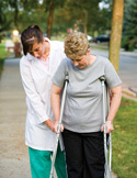 A healthcare worker in a white coat assists an elderly woman practicing walking with crutches on a sidewalk.