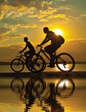 Silhouettes of two people riding bicycles on a beach at sunset with a glowing sun and water reflection.