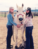 Two women stand on either side of a white horse in a dirt paddock, posing with their hands on the horse.