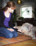 A woman kneeling on a tile floor gently interacting with a fluffy grey dog.