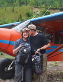 A man kissing a smiling woman on the forehead in front of a small orange and blue bush plane.