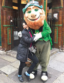 A woman happily hugging a person in a large leprechaun mascot costume on a city sidewalk.
