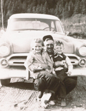 Vintage black and white photo of a man sitting on the bumper of an old car holding two small children.
