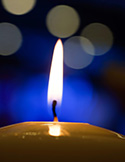 Close-up of a single lit candle flame against a soft blue background with blurred white bokeh lights.