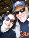 A smiling man and woman wearing sunglasses and baseball gear at a stadium.