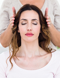 A woman with eyes closed receiving Reiki as a practitioner’s hands hover near the sides of her head.