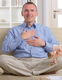 A man sitting cross-legged on a sofa with his hands placed over his heart in a self-Reiki healing position.