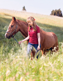 A woman in a red shirt walks a brown horse through a sunlit, grassy field with a hillside in the background.
