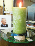 A lit green pillar candle on a glass plate with stones and a portrait of an elderly man in the background.