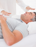 A Reiki practitioner holds their hands over a man lying down with his eyes closed during a healing session.