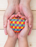 Larger hands cradling smaller hands that hold a heart patterned with colorful puzzle pieces.