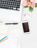 Top-down view of an organized white desk with a laptop, smartphone, pink flowers, and stationery.