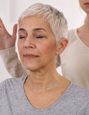A woman with short grey hair keeps her eyes closed while a practitioner holds hands near her head.