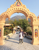 A woman standing before a decorative stone archway leading to a building with mountains in the background.