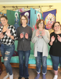 Four women standing in a row indoors, smiling and holding their hands up in a Reiki healing position.