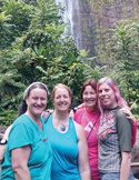 Four women smiling and hugging outdoors in front of a lush green forest and a tall waterfall.