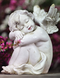 A small, white stone statue of a sleeping angel cherub nestled among pink flowers and green leaves.