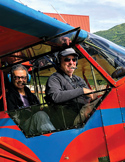 Two men smiling and wearing headsets inside the cockpit of a bright red and blue small aircraft.