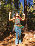 A woman in a colorful fringed top dances with arms raised in a sunlit forest.