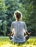 Back view of a woman meditating in a cross-legged position on grass in a sunlit park.