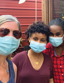 Three smiling women wearing light blue surgical face masks pose closely together outdoors.