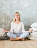 Young woman sitting in a meditative cross-legged pose with eyes closed in a bright room.