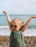 A happy toddler wearing sunglasses with arms raised joyfully on a pebble beach.