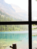 View of a serene turquoise lake and misty mountains through a dark wooden window frame.