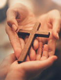 Two hands gently cradling a small, simple wooden cross in a warm, soft-focus setting.