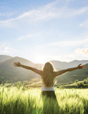 Woman standing in a sunny green field with her arms outstretched toward mountains and a bright sky.