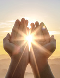 Two hands cupped toward the sky with bright sunlight bursting through the center against a mountain horizon.