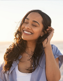 A woman with long curly hair smiles brightly with her eyes closed, basking in warm sunlight at the beach.