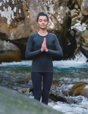 A woman stands in a meditative prayer pose with her eyes closed in front of a rushing mountain river.