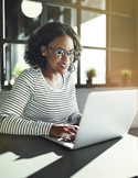 A smiling woman with glasses working on a laptop in a bright, modern office space.