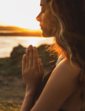 Side profile of a woman with eyes closed and hands in prayer position against a sunset over water.