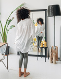 A woman with curly hair looks at her reflection in a large floor mirror in a bright room.