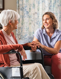 A smiling volunteer sits and holds the hand of an elderly woman in a wheelchair.