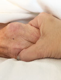 A close-up of two hands gently holding each other on white bedding, showing comfort and connection.