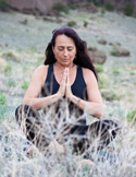 A woman meditating with hands in prayer position in a rocky, natural outdoor landscape.