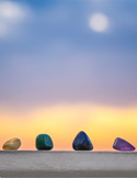 Four colorful tumbled gemstones sitting on a ledge against a soft blue and orange sunset sky.