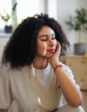 A woman with curly hair sits peacefully with eyes closed in a bright, sunlit room.
