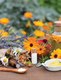 A collection of herbal remedies, including fresh marigolds, lavender, oils, and powders on a wooden table.