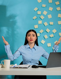 A stressed woman at an office desk with her hands raised, surrounded by a wall covered in yellow sticky notes.
