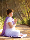 A woman in a purple shawl sitting in a meditative prayer pose on a sunlit forest path.