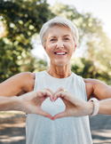 A smiling senior woman in a white tank top making a heart shape with her hands outdoors.