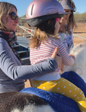 A young child wearing a pink helmet is assisted by an adult while sitting on a pony.
